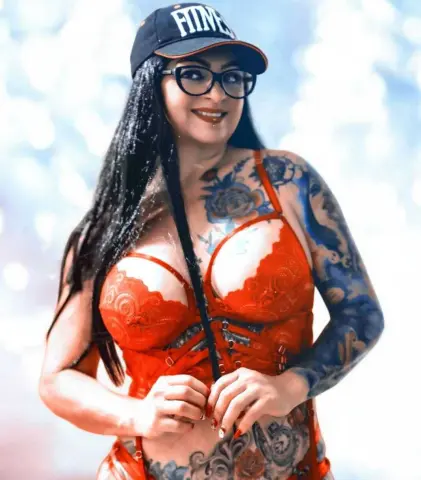 Smiling tattooed woman in red lace lingerie and a black baseball cap posing outdoors in bright sunlight.