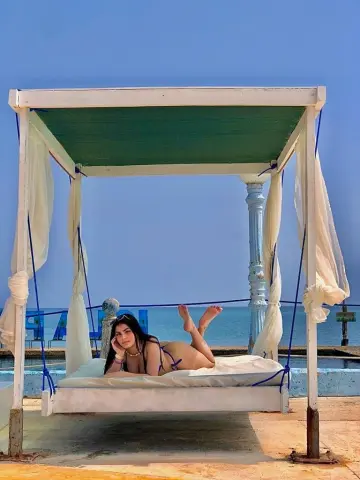 Young woman in a blue bikini relaxing on a white cabana bed by the ocean under a clear blue sky.