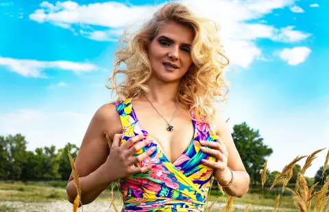 Blonde woman in a colorful floral summer dress posing in a sunny field under a blue sky.