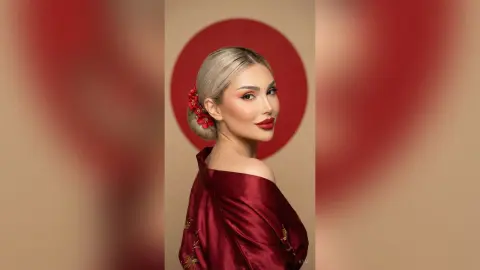 Elegant woman in a red satin gown with blonde hair and red floral accessories posing against a gold backdrop.