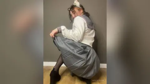 Young woman in a white sailor blouse and grey pleated school skirt posing in a minimalist indoor setting.