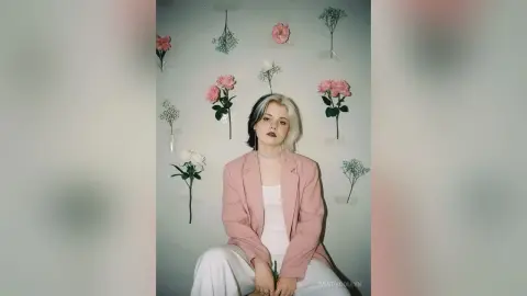 Blonde woman in a pink blazer and white trousers posing against a floral wall background with a dreamy mood.