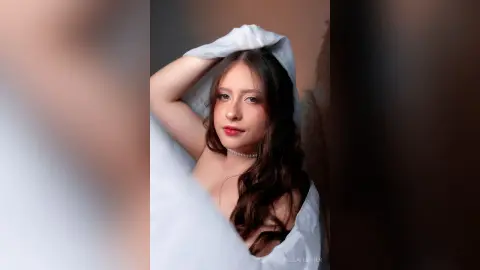 Young woman with long wavy hair wearing a white garment posing gracefully in a soft, dreamy studio setting.
