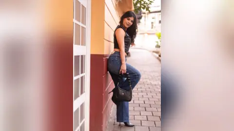 Smiling woman in a black sleeveless top and blue jeans leaning against a colorful building on a sunny street.
