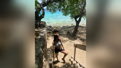 Smiling woman in a blue bikini sitting on wooden stairs by a tropical turquoise ocean beach.