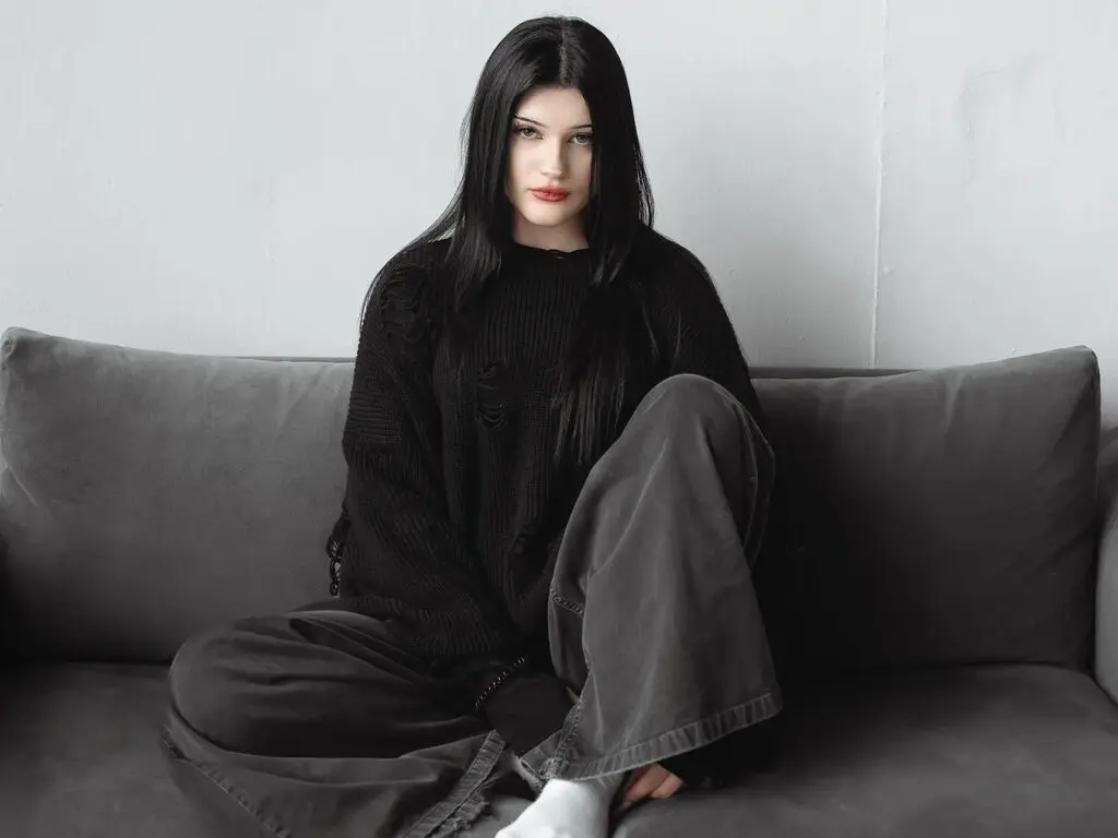 Moody portrait of a young woman with long black hair wearing a black sweater and grey trousers on a grey sofa.