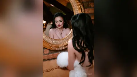 Glamorous woman with long dark hair in a white faux fur wrap looking into a vintage gold mirror.
