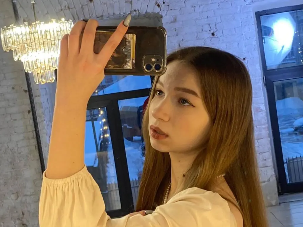 MagalyBoffa takes a mirror selfie wearing a light cream blouse, showing her long brown hair and light makeup.