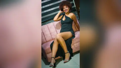 Smiling woman in a chic black evening gown posing on a pink velvet sofa in a modern living room.