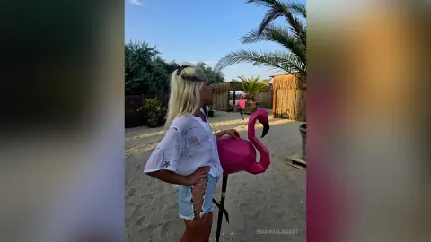 Blonde woman in a white linen shirt and denim shorts standing by a pink flamingo statue on a sunny beach.