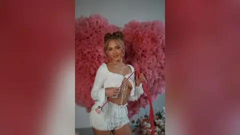 StellaFletcher wears a white lacey ruffled top and matching shorts while posing in front of a large pink heart-shaped ruffly backdrop.