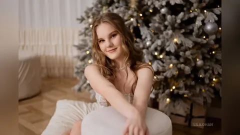 Young woman in a white dress sitting by a glowing Christmas tree in a cozy festive setting.