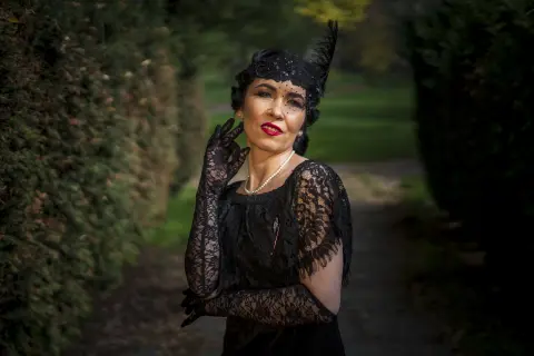 Elegant woman in vintage black lace outfit and pearl necklace posing in a moody garden setting.