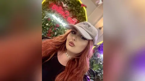 Redheaded woman in a black top and grey baseball cap posing in a trendy neon-lit floral setting.