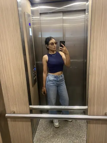 Stylish woman in a navy blue crop top and light wash jeans taking a mirror selfie in a modern elevator.