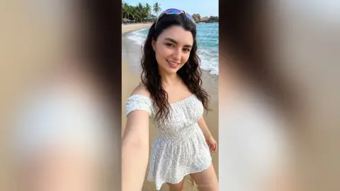 Smiling young woman in a white floral sundress enjoying a sunny day at a tropical beach.