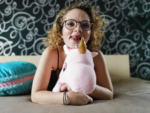 Smiling curly-haired woman in a black top holding a pink unicorn plushie while relaxing on a couch.