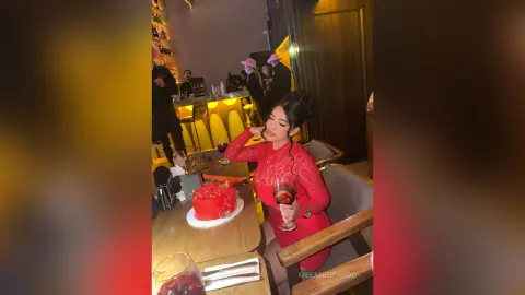 Elegant woman in a red dress sitting at a restaurant table with a red cake in a warm, moody setting.