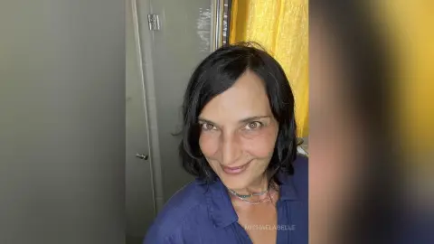 MichaelaBelle smiles while wearing a blue button-down shirt, showing her dark hair and light eyes.