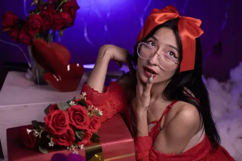 CloeLeee wears a red lace teddy and a large red bow in her hair while posing next to red roses and a heart-shaped bowl.
