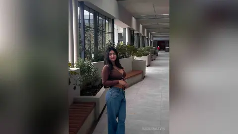 Young woman in a brown long-sleeve top and blue jeans standing in a modern outdoor hallway with plants.