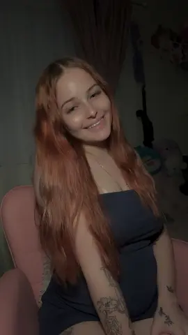 Smiling young woman with long strawberry blonde hair wearing a navy blue dress in a cozy indoor setting.