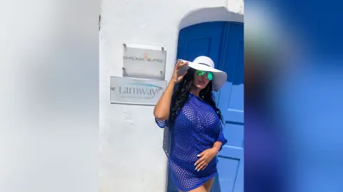 MissyJolie stands against a white wall wearing a blue mesh dress and a white sun hat.