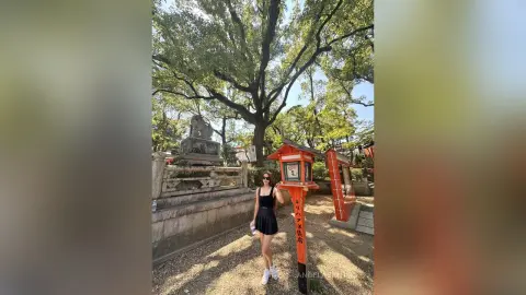 Woman in a black summer dress and sunglasses standing by a traditional red shrine under lush green trees.