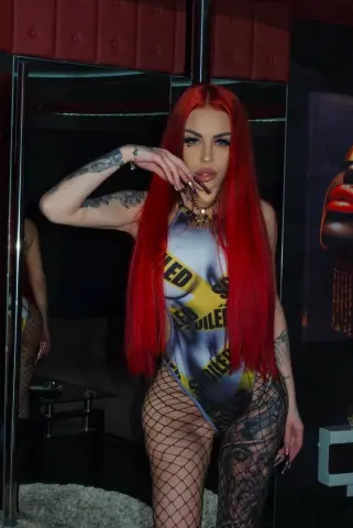 NaomyBlackBarb poses in a blue and yellow bodysuit and fishnet tights, showing off her long red hair and arm tattoos.