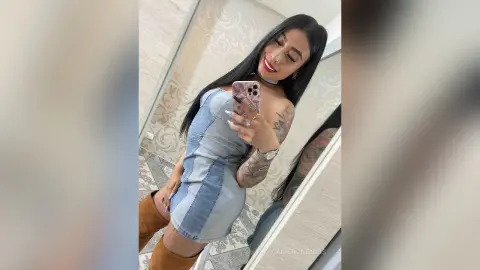 Smiling woman in a denim mini dress and brown suede boots taking a mirror selfie in a bright bathroom.