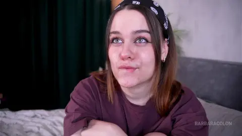 Young woman in a purple t-shirt smiling thoughtfully in a cozy indoor setting.