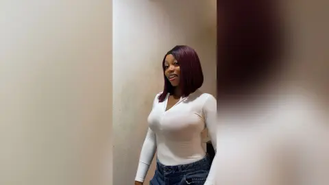 Smiling young woman in a white long-sleeve bodysuit and blue jeans standing against a neutral wall.
