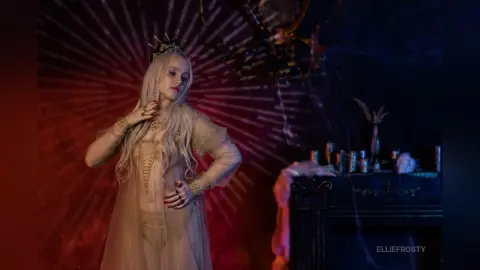 EllieFrosty poses in a sheer, long-sleeved gold gown and a crown, standing against a red sunburst background.