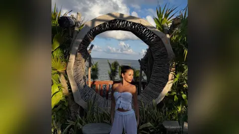Woman in white summer outfit standing by a rustic wooden archway overlooking the ocean at sunset.