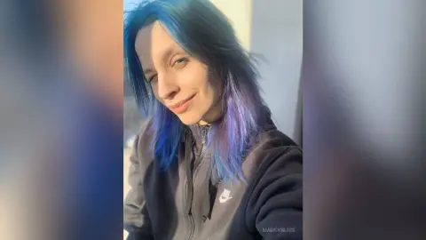 Smiling woman with blue hair wearing a black Nike hoodie in bright natural sunlight.