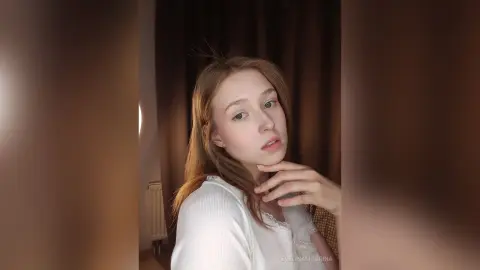 Young woman in a white lace top posing with a soft, dreamy expression in a warm indoor setting.
