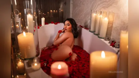 Nude woman relaxing in a white bathtub filled with red rose petals surrounded by glowing candles in a romantic setting.