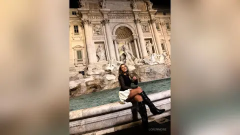 Stylish woman in a black top and white skirt sitting by the Trevi Fountain in Rome at night.