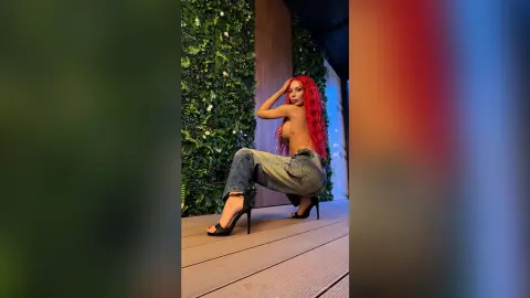 Red-haired woman in denim jeans and black high heels posing on a wooden deck against a lush green wall.
