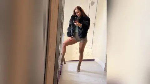 Stylish woman in a black faux fur jacket and patterned shorts taking a mirror selfie in a bright room.