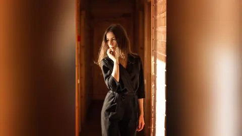 Thoughtful woman in a black jumpsuit standing in a sunlit rustic wooden hallway with warm moody lighting.