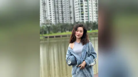 Young woman in a denim jacket and grey tank top posing by a calm lake near city buildings.