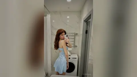 Young woman in a light blue slip dress taking a mirror selfie in a modern marble bathroom.