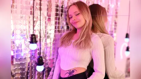 LucyLoy smiles with her eyes closed, wearing a white long-sleeve crop top and showing a spider tattoo on her stomach.