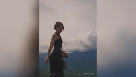 RossiNoleen stands outdoors against a cloudy sky wearing a black crop top and black leggings, showing off her arm tattoos.