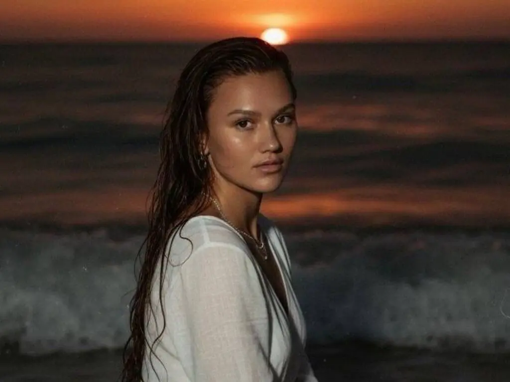 Young woman in a white linen shirt at the beach during a moody sunset over the ocean waves.