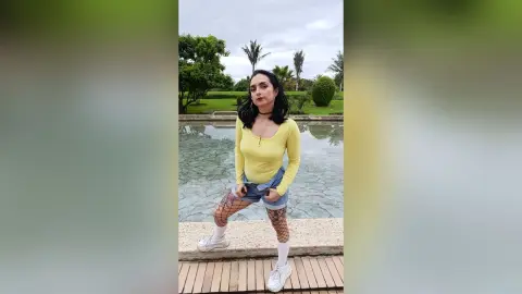 Young woman in a yellow long-sleeve top and denim shorts posing by a serene outdoor pond.