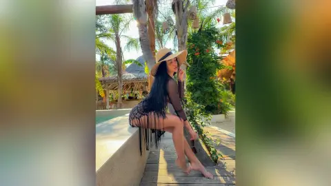 NicoleBella sits on a stone ledge wearing a black sheer mesh bodysuit with long fringe and a wide-brimmed straw hat.