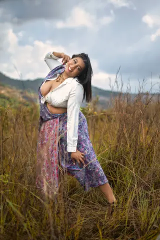 Smiling woman in a white blouse and patterned long skirt posing in a grassy field under a cloudy sky.