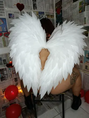 Woman in white angel wings and crown sitting amidst red balloons and newspaper collage decor.